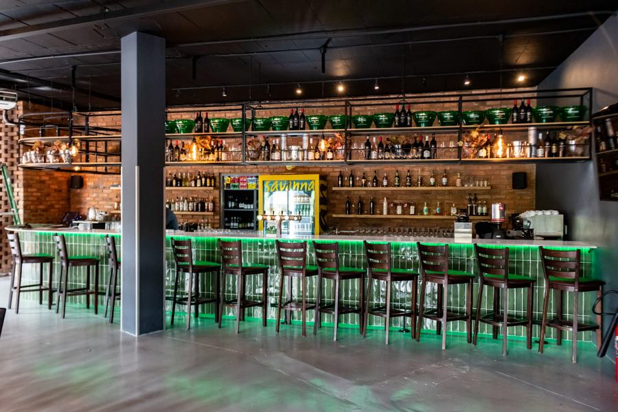 Long bar with a green glazed tile front, under-counter lighting and steel shelving against a brick wall